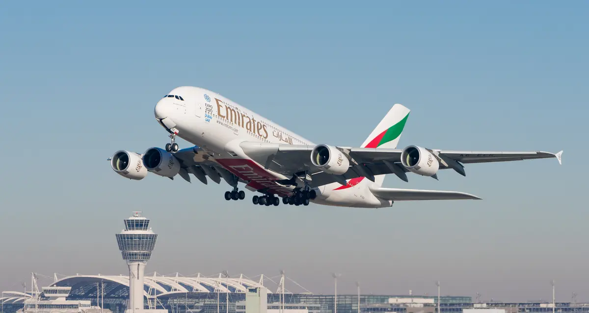 Um Airbus A380 da Emirates recém-saído do solo, com a torre de controle e o terminal de um aeroporto ao fundo.