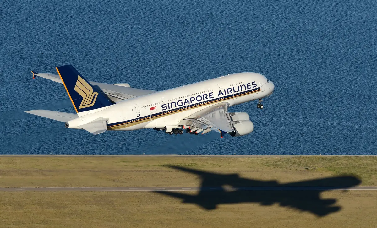 Um Airbus A380 da Singapore Airlines decolando sobre a água, com a sombra se estendendo pela grama abaixo.