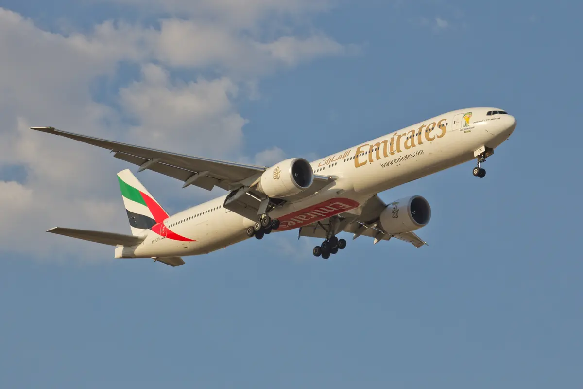 Um Boeing 777-300ER da Emirates em voo contra um céu azul com algumas nuvens, com o trem de pouso sendo recolhido.